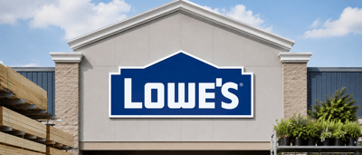 Lowe's storefront exterior with the company logo sign, stacked lumber, and potted plants displayed outside.
