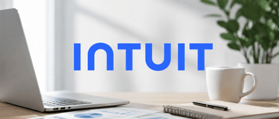Intuit logo displayed over a desk with a laptop, coffee mug, notebook, plant, and financial chart printout.