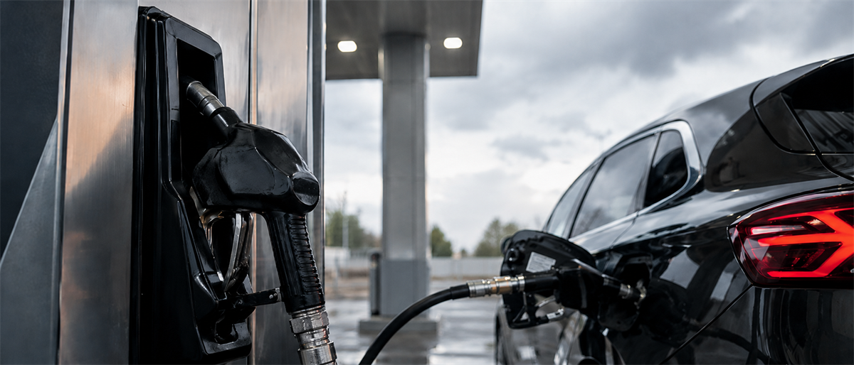 A fuel pump nozzle refuels a black car at a gas station under an overcast sky.