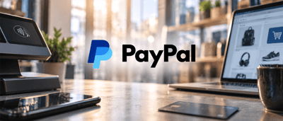 PayPal logo displayed on a desk with a payment terminal, smartphone, laptop showing an online shopping page, and a credit card.