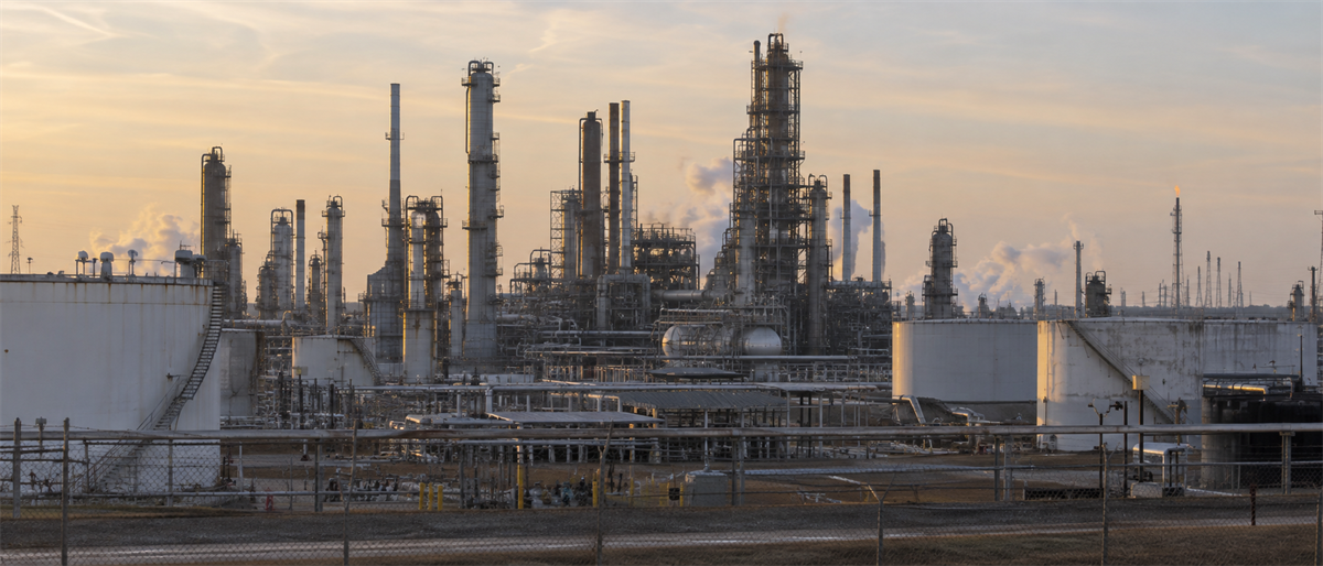 Oil refinery complex with storage tanks, distillation towers, and piping, photographed at dusk with a chain-link fence in foreground.