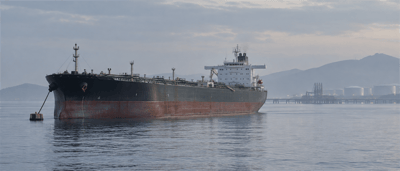 An oil tanker anchored near a port terminal with storage tanks and hills visible in the background.