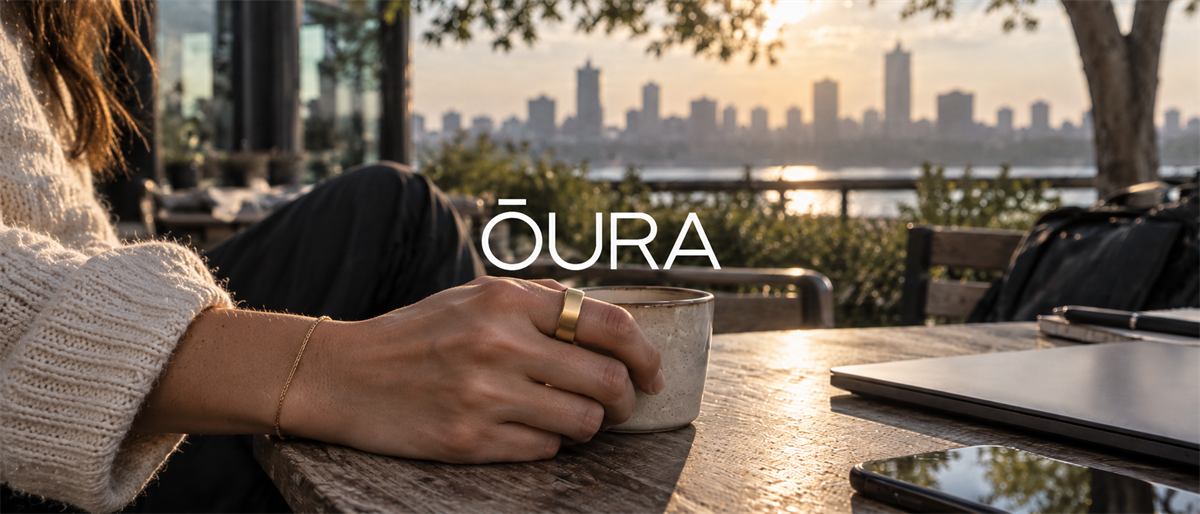 Woman wearing an Oura ring holds a coffee cup at an outdoor table with a city skyline behind her.