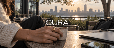 Woman wearing an Oura ring holds a coffee cup at an outdoor table with a city skyline behind her.