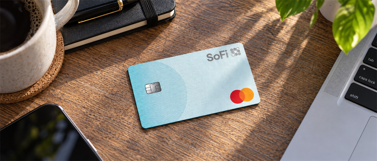 A SoFi debit card with Mastercard logo rests on a wooden desk near a coffee mug and laptop.