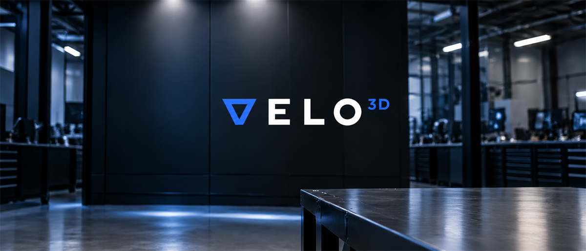 Velo3D logo illuminated on a dark wall inside an industrial manufacturing facility with equipment visible.