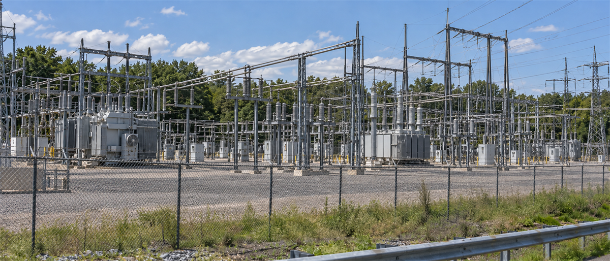 Electrical substation with transformers, transmission towers, and power lines behind a chain-link fence surrounded by trees.