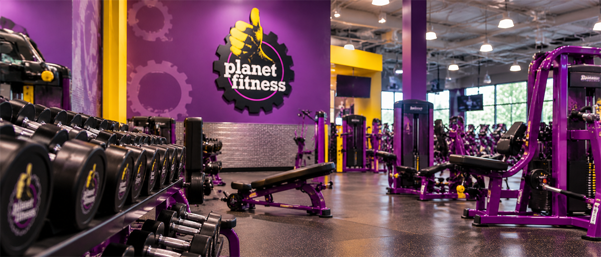 Interior of a Planet Fitness gym showing the brand logo, dumbbell rack, benches, and purple exercise equipment.