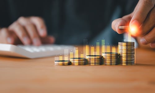 Hand placing a coin on stacked columns of coins with a rising bar chart overlay, representing dividend growth and income investing.