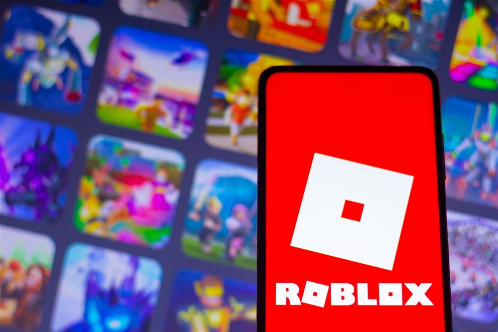 roblox logo on smartphone with blurred images on computer screen in background