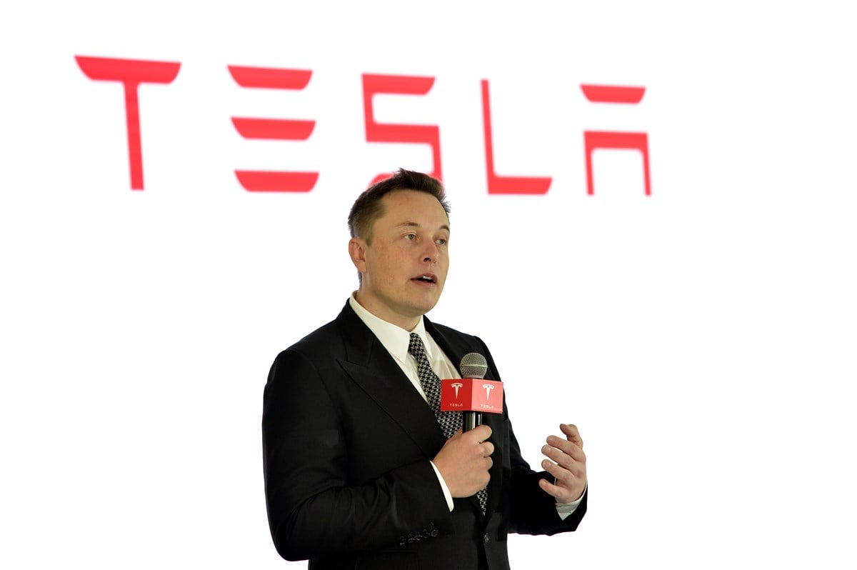 photo of elon musk holding microphone with tesla banner and logo in background