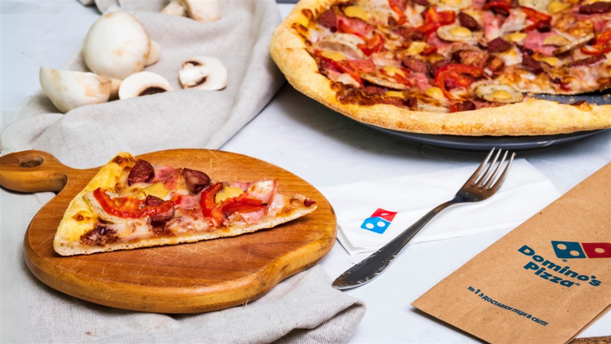 A slice of Dominos pizza on a wodden cutting board next to a full pie