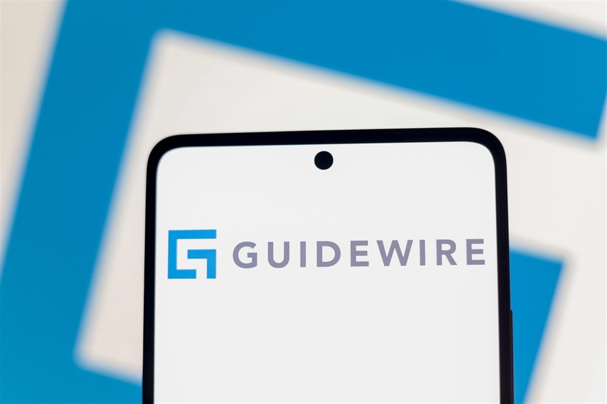 Guidewire logo displayed on a smartphone screen against a blue geometric backdrop, highlighting momentum in AI-driven software adoption and improving profitability.