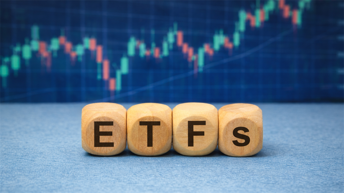 Wooden blocks spelling “ETFs” on a light blue surface, with a blurred candlestick stock chart on a screen behind.