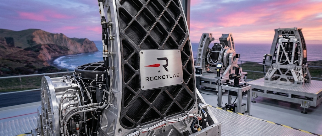 Rocket Lab-branded aerospace hardware on an outdoor platform with a coastal backdrop at sunset.