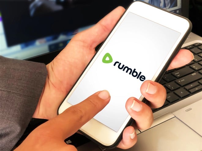 Man using video application Rumble on mobile phone with laptop in the backdrop.
