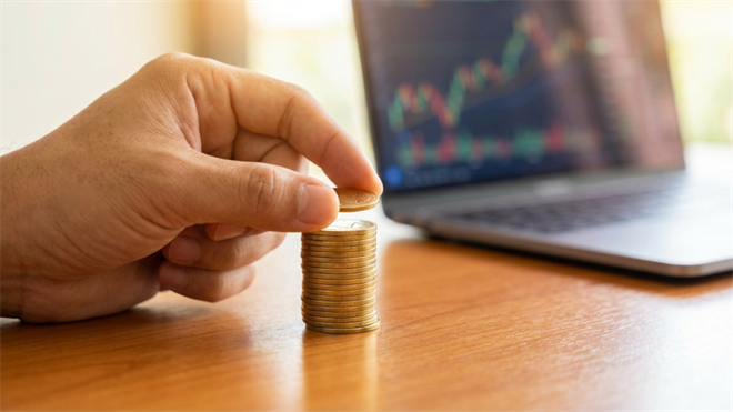 Hand stacking coins beside rising stock chart on laptop, symbolizing steady gains from tollbooth-style investment stocks.