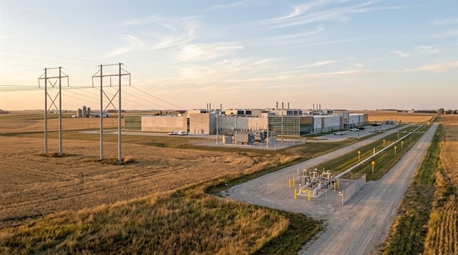 An aerial view of a large modern facility, likely a data center, situated among flat agricultural fields at sunset, flanked by power lines and utility infrastructure.