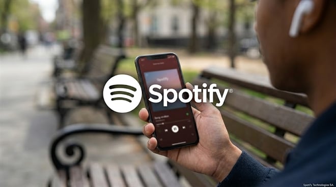 A person wearing wireless earbuds holds a smartphone displaying the Spotify app outdoors.