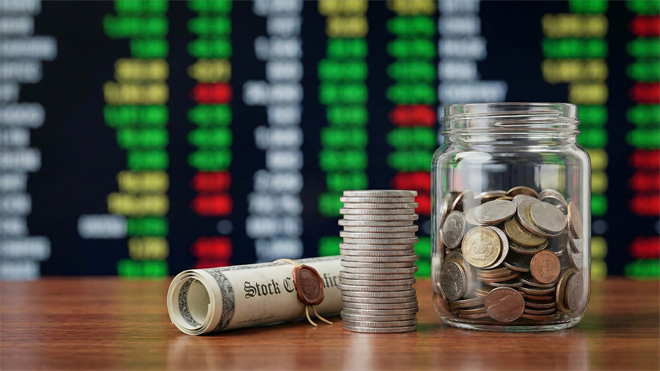 A rolled stock certificate, a stack of coins, and a jar of coins sit before a blurred stock ticker board.