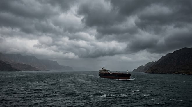 A cargo tanker ship navigates choppy open waters under a dark, overcast sky near rocky coastlines.