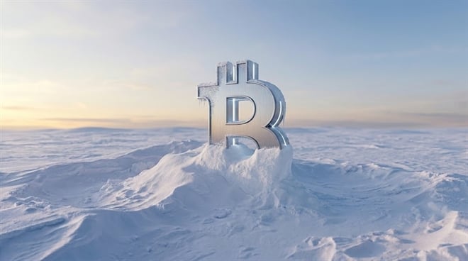 A metallic Bitcoin symbol partially buried in snow against a pale winter sky.