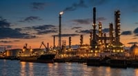 An illuminated oil refinery and docked tanker ship beside water at sunset.