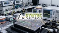 Syntec Optics logo overlaid on a photonics laboratory workbench with optical testing equipment and circuitry.