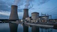 A nuclear power plant with two large cooling towers emitting steam, reactor buildings, and a waterway in the foreground.