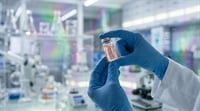 A gloved researcher holds a small glass vial containing a pink liquid in a laboratory setting.
