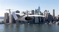 A white electric vertical takeoff and landing aircraft flies over a city skyline and waterway.