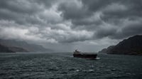 A cargo tanker ship navigates choppy open waters under a dark, overcast sky near rocky coastlines.