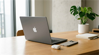 An Apple MacBook, iPhone, and AirPods case sit on a wooden desk beside a notebook.