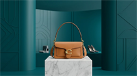 A tan leather Coach shoulder bag with gold hardware displayed on a marble pedestal.