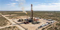 An aerial view of an active oil drilling rig in a vast, arid desert landscape with support vehicles, storage tanks, and a small gas flare under a blue sky.