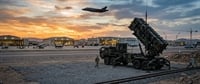 A Patriot missile launcher and soldier on an airbase tarmac as an F-35 fighter jet flies overhead at sunset.
