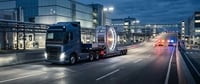 A semi-truck hauls a large cylindrical industrial component past police cars on a nighttime industrial roadway.