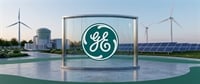 General Electric (GE) logo displayed on glass panel with wind turbines and solar panels in the background.