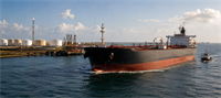 Large oil tanker ship departing a port terminal with storage tanks and cranes visible near the water.