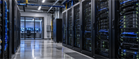 Data center hallway with rows of server racks and cables, plus a monitoring room with computer screens in the background.