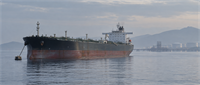 An oil tanker anchored near a port terminal with storage tanks and hills visible in the background.