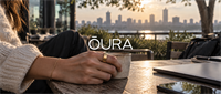 Woman wearing an Oura ring holds a coffee cup at an outdoor table with a city skyline behind her.