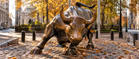 The Charging Bull bronze statue near Wall Street, surrounded by fallen autumn leaves and city buildings.