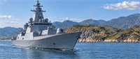 Gray naval warship equipped with radar and weapons systems sails through coastal waters near mountainous terrain.