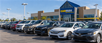 Rows of used vehicles parked in a lot outside a CarMax dealership building with its logo displayed.