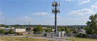 A cellular communications tower with multiple antennas stands behind a fenced equipment enclosure in a suburban area.