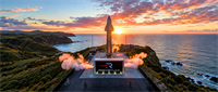 Rocket Lab rocket stands on a coastal launch pad with vapor clouds and the Rocket Lab logo visible at sunset.